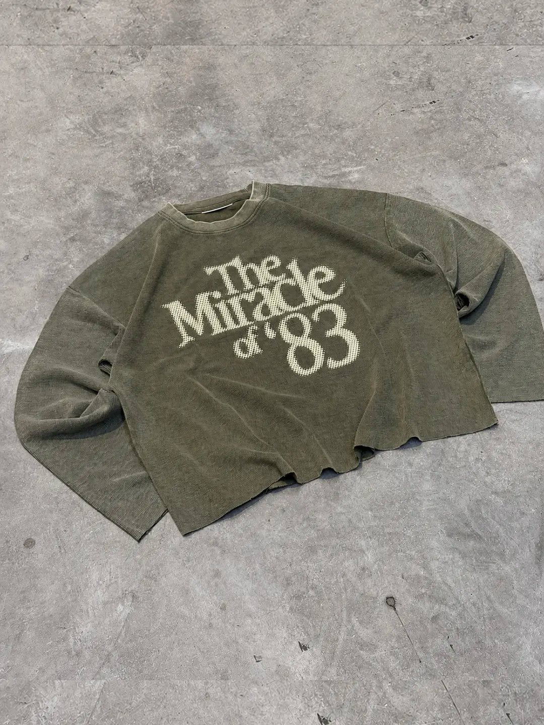 An olive green long-sleeve t-shirt with a distressed cream graphic reading 'The Miracle of '83', laid flat on a textured grey concrete surface.