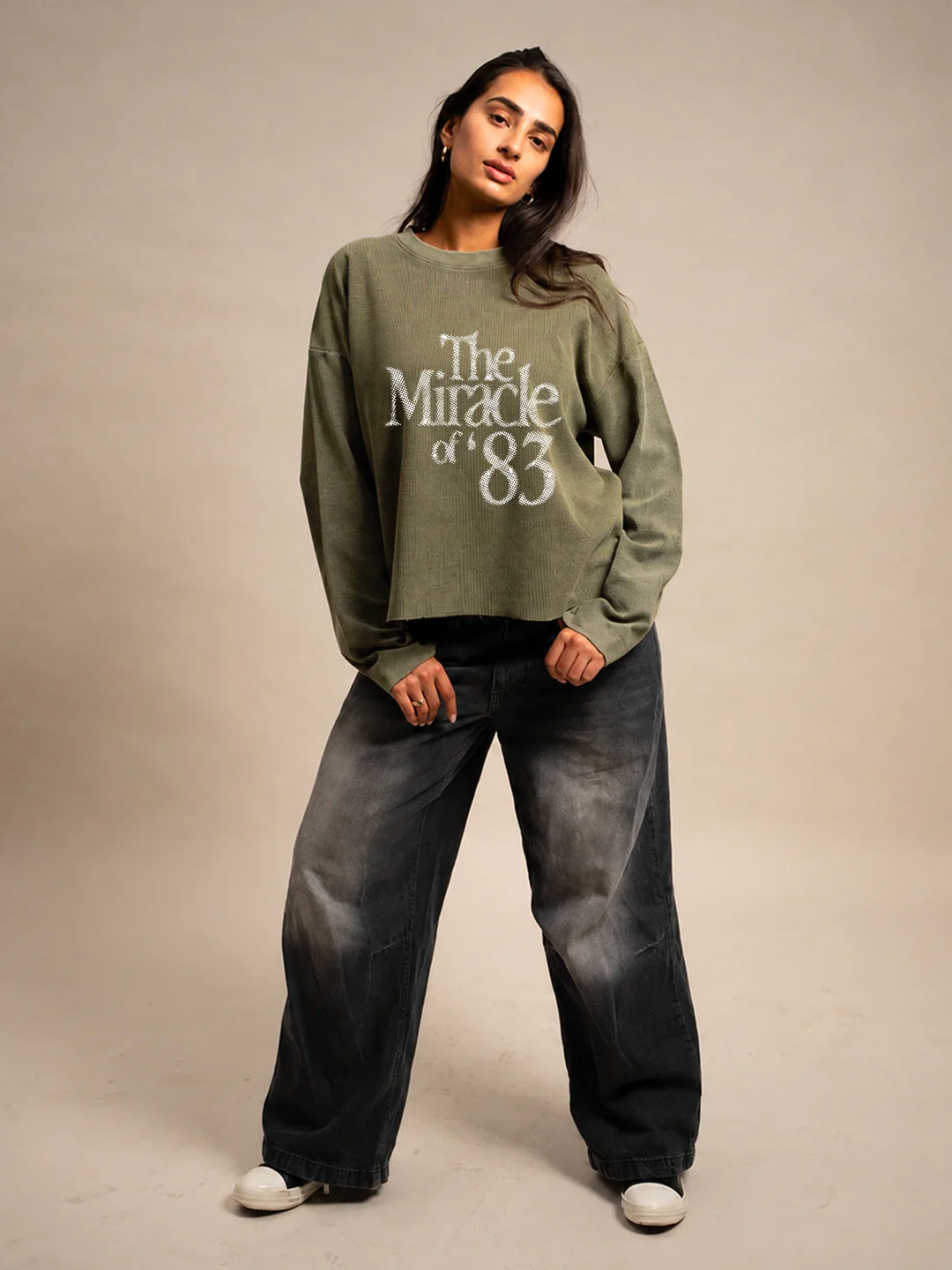 A woman modeling a cropped sage green t-shirt with 'The Miracle of '83' printed in white across the front, paired with wide-leg faded black jeans and white sneakers, against a plain beige studio background.