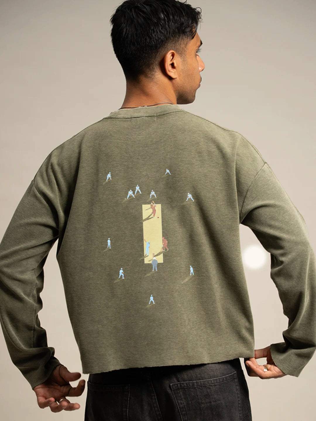A man wearing an olive green long-sleeve t-shirt with a graphic print on the back, paired with dark jeans, against a neutral studio background.