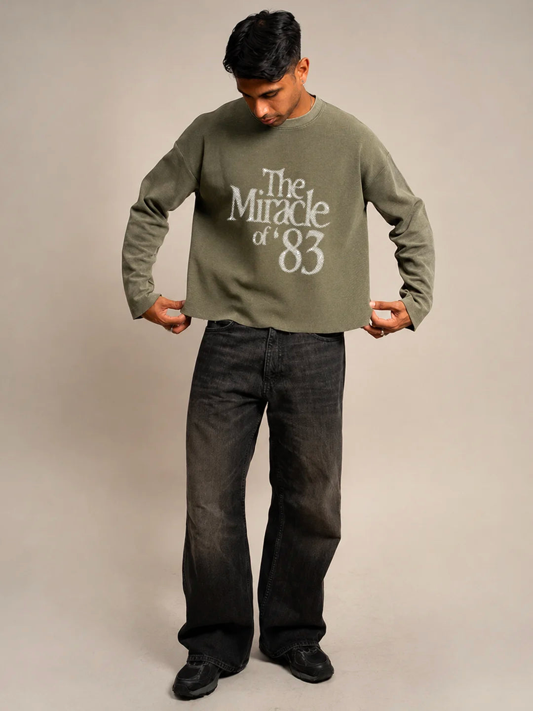 A man wearing an olive green long-sleeve crewneck t-shirt with a distressed white graphic reading 'The Miracle of '83' on the front, paired with dark grey jeans