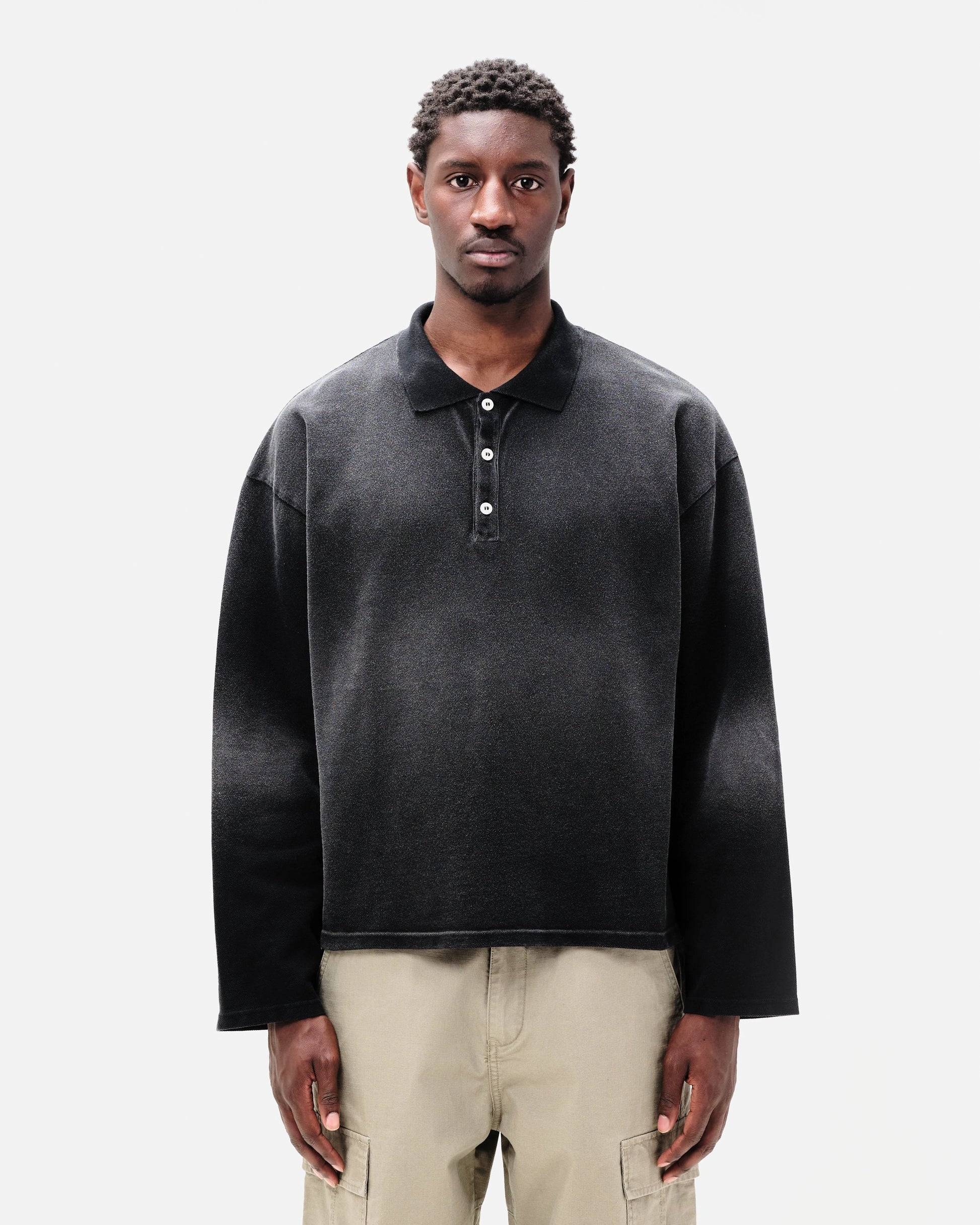 Man wearing a dark grey oversized polo shirt with beige pants on a white background