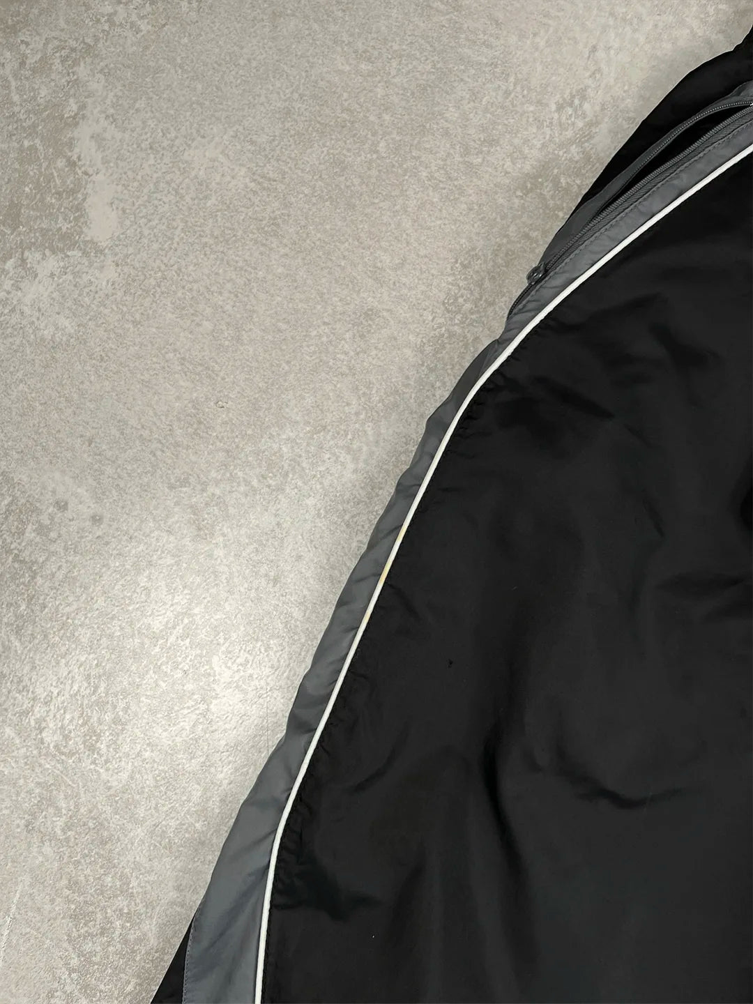 Close-up of a black garment laid flat on a light grey textured surface, showing a dark grey side panel and a thin white piping detail.