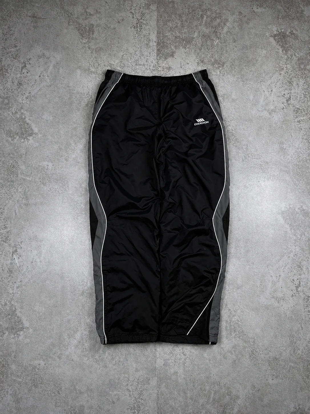 Black athletic pants with grey side panels and white piping details, laid flat on a textured grey background.