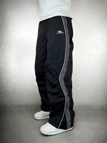 Black track pants with a grey side stripe and white logo, worn by a model with white sneakers against a textured grey wall.
