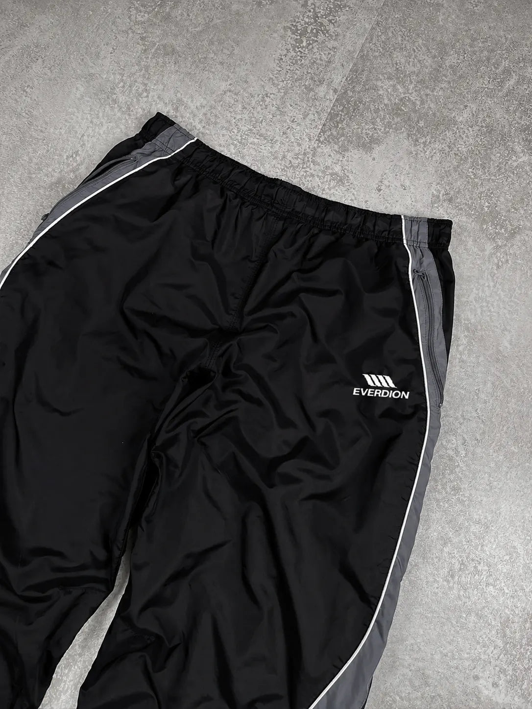 Black Everdion athletic windbreaker pants with grey side stripes and a white logo, laid flat on a grey concrete floor.