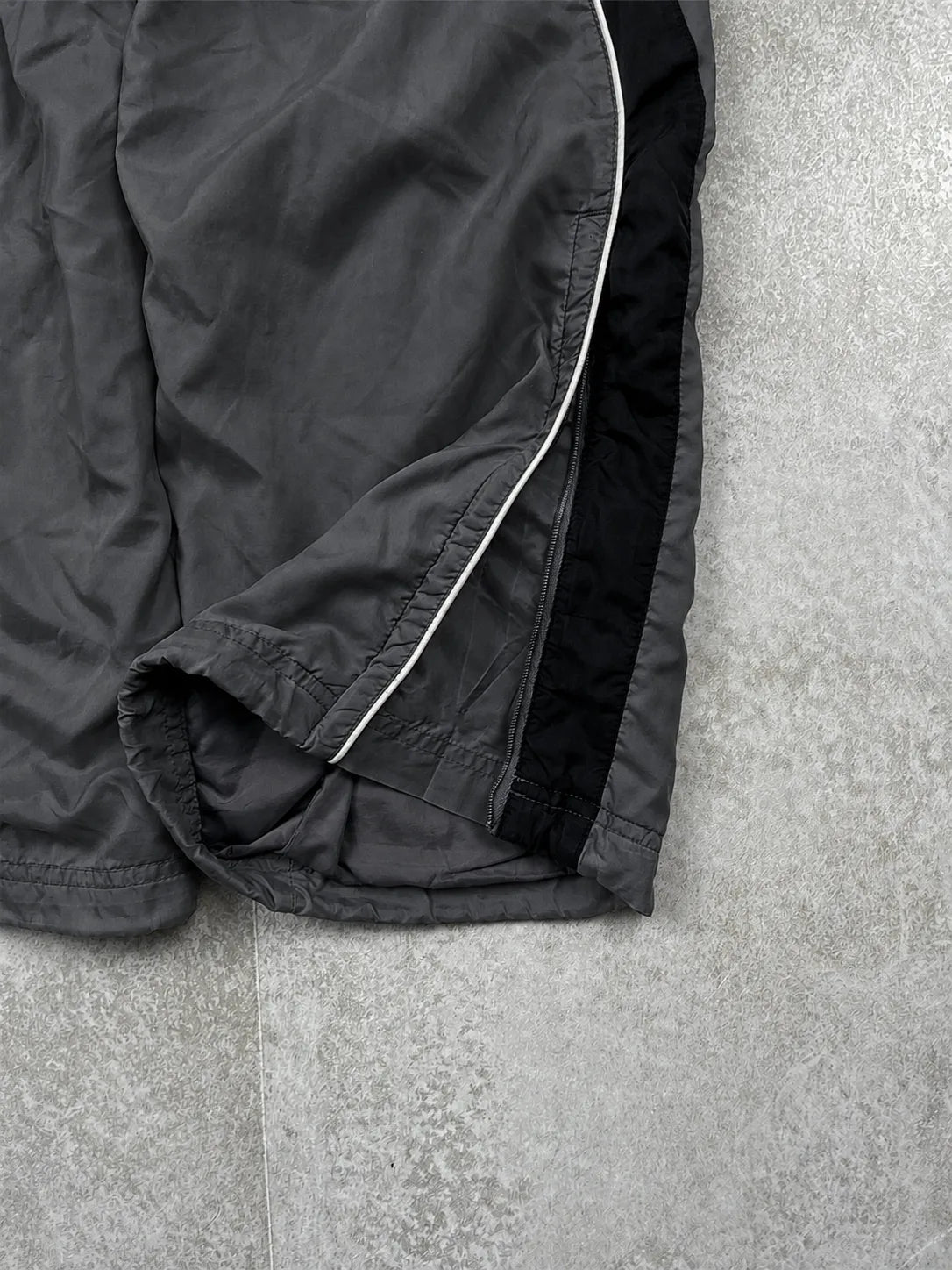Close-up of dark grey athletic shorts laid flat on a light grey textured surface, showing a black side panel with a thin white piping detail.