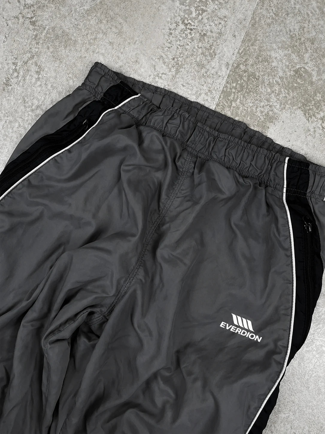 Dark grey Everdion athletic track pants with black side panels and white piping details, laid flat on a grey textured floor.