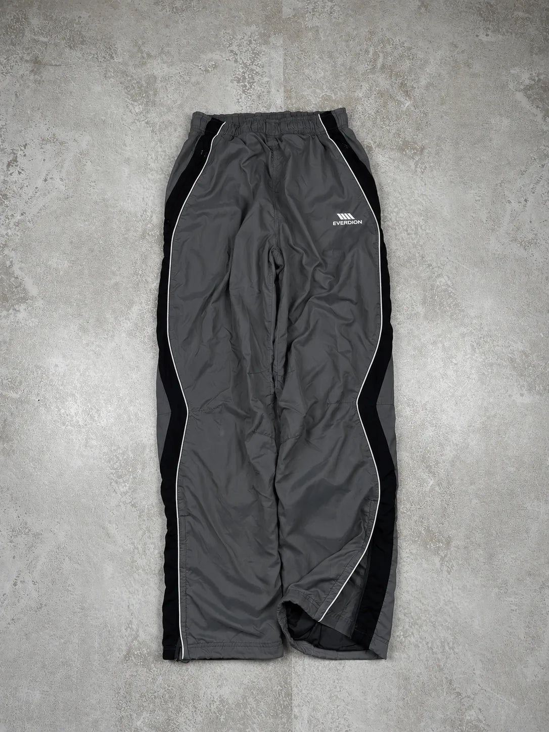 Grey and black athletic pants with white side stripes and a white embroidered logo on the left thigh, laid flat on a textured grey concrete surface.