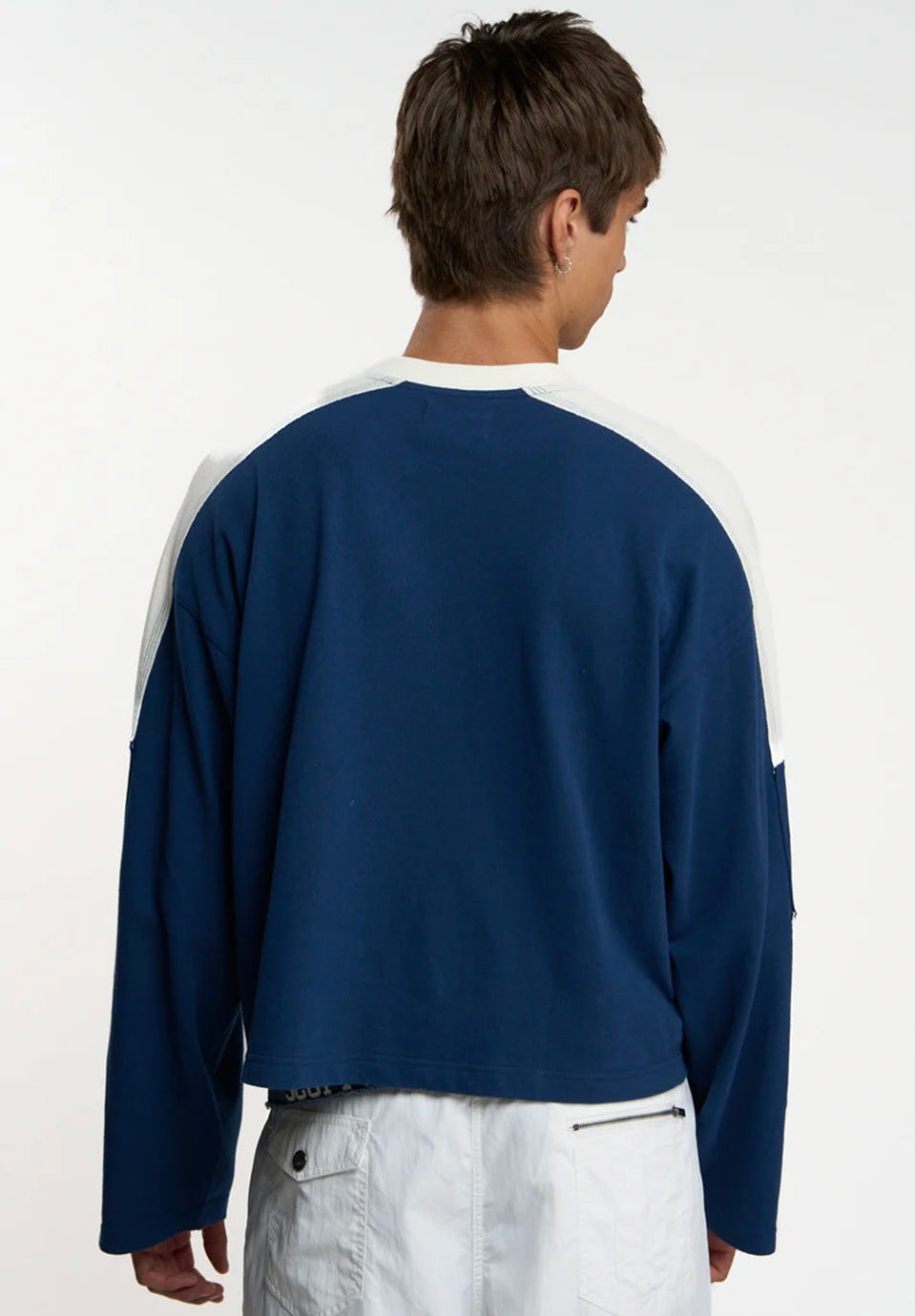 A man viewed from behind wearing a navy blue long-sleeve crop t-shirt with white color-blocked shoulder panels and white pants, set against a plain white studio background.