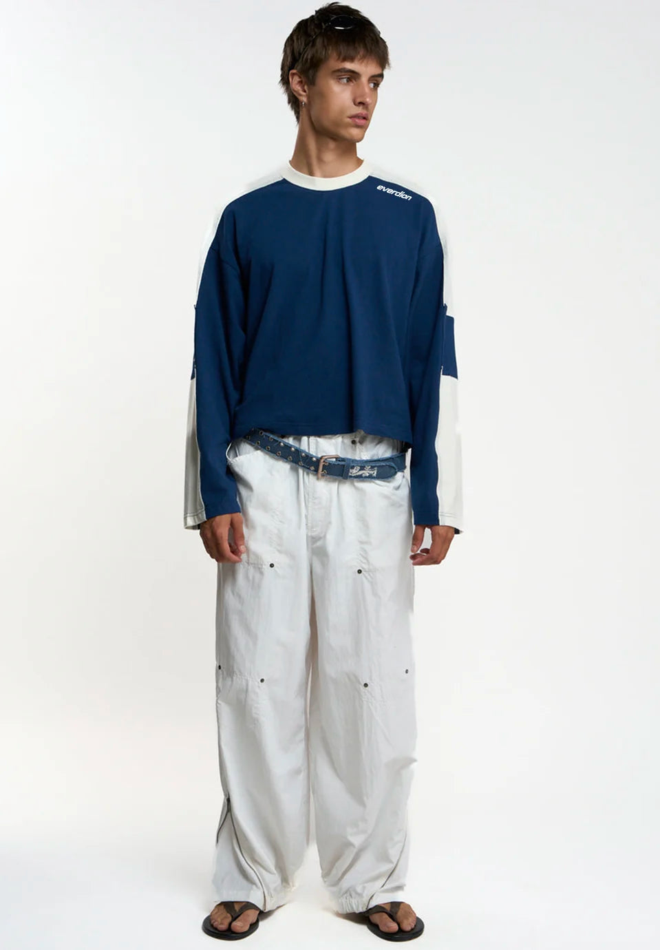 A man wearing a navy blue and white colorblock long-sleeve t-shirt with 'everdion' printed on the chest, paired with white pants and a denim belt, against a plain white studio background.