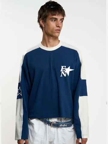 A man wearing a navy blue and white colorblock long-sleeve shirt with a white star logo on the chest, paired with white pants and a denim belt, against a plain white studio background.