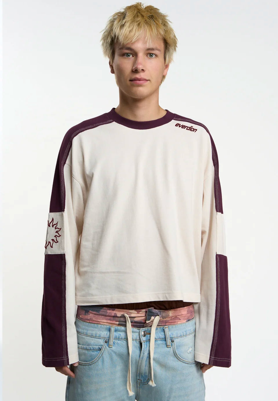 A model wearing a cream and dark purple long-sleeve crop top, light blue distressed jeans with Everdion logo