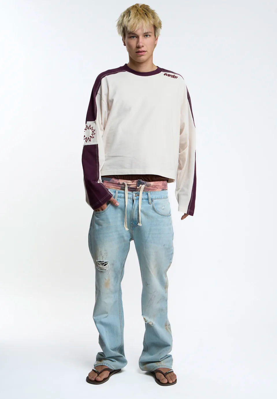 A model stands against a white studio background wearing a cream and dark purple long-sleeve crop top, light blue distressed jeans, and brown sandals.
