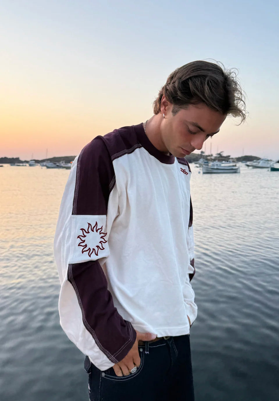 A man wearing a white and dark plum raglan long-sleeve t-shirt with a flame graphic on the sleeve, standing by a calm harbor at sunset.