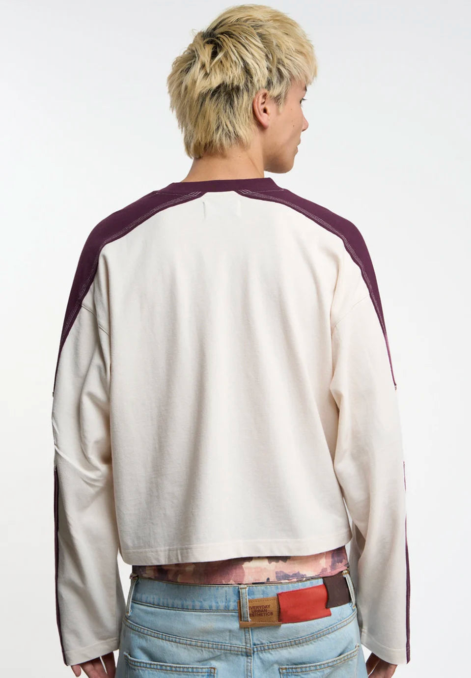 A person wearing a cropped long-sleeve t-shirt with an off-white body and dark plum raglan sleeves, paired with light blue jeans and a patterned belt, against a plain white studio background.