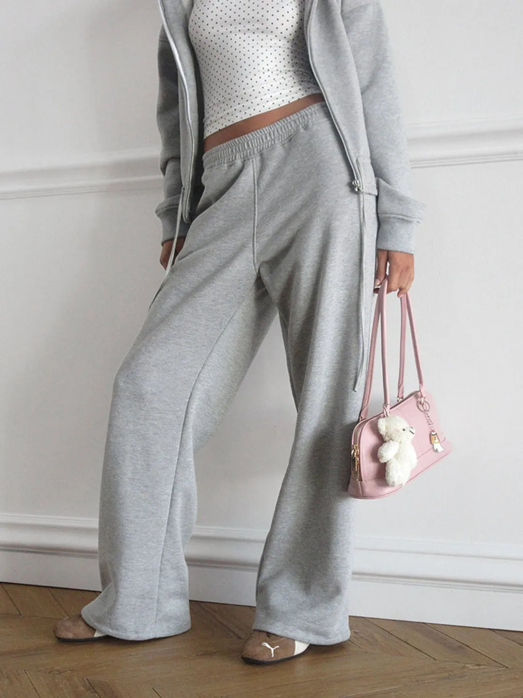 Person wearing a gray tracksuit holding a pink handbag with a teddy bear on a wooden floor.