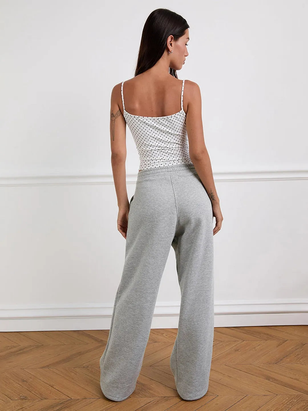Woman wearing a white patterned top and gray sweatpants against a white wall.