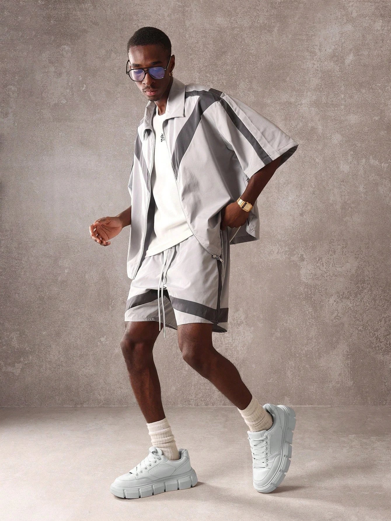 Man wearing a stylish outfit with a patterned shirt and shorts against a grey background