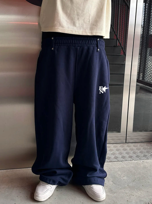 A person wearing navy blue wide-leg sweatpants with a white embroidered logo on the left thigh, paired with a cream t-shirt and white sneakers, standing in front of a metallic door.