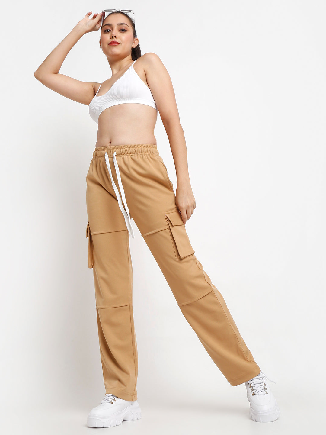 A female model wearing tan utility cargo pants with large side flap pockets and a drawstring waistband, paired with a white cropped tank top.