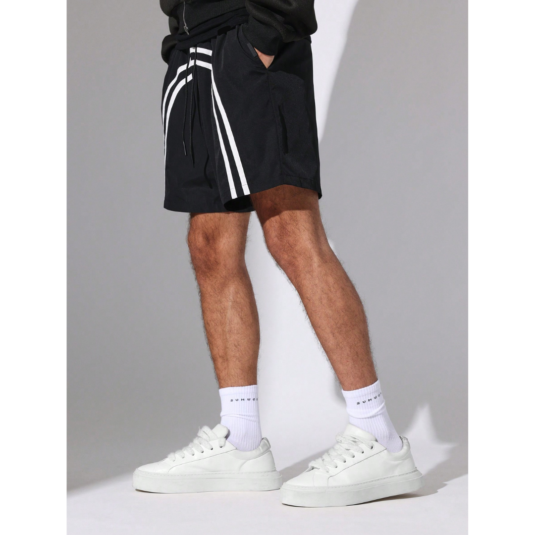 Person wearing black shorts with white stripes and white sneakers on a grey background