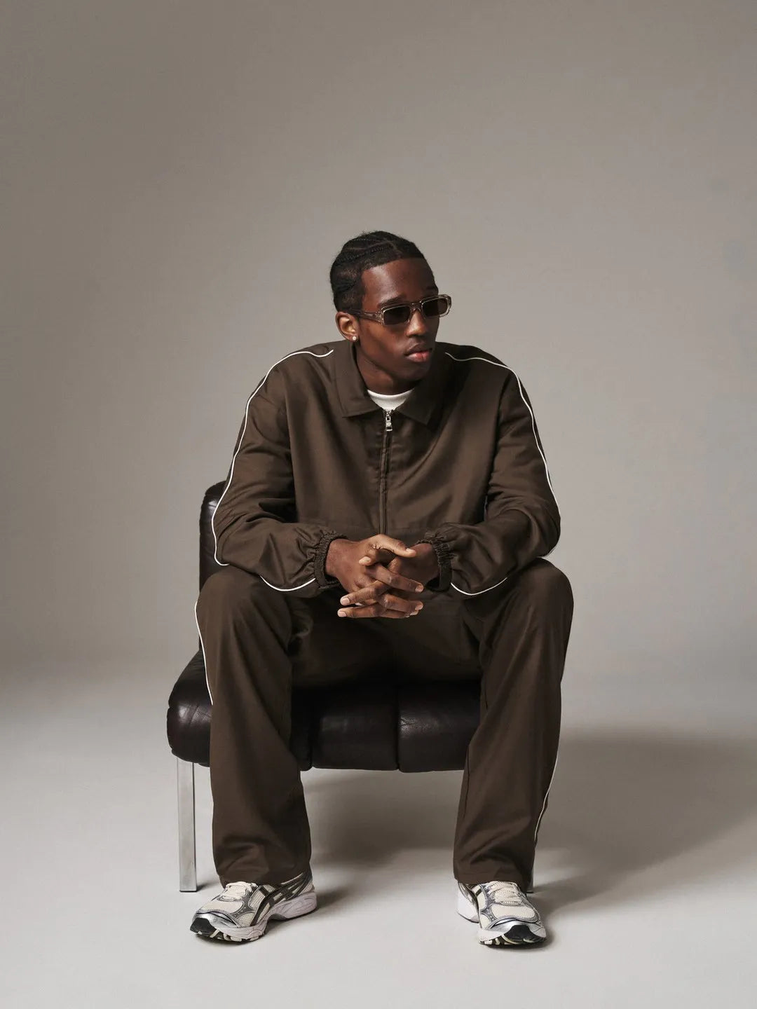 Man in a brown tracksuit sitting on a black chair against a plain background