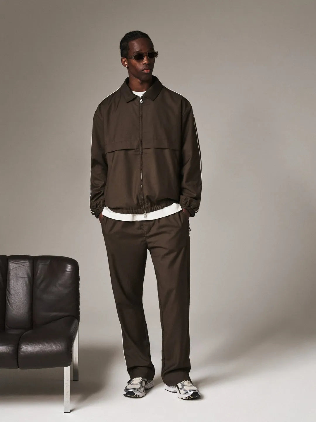 Man wearing a brown tracksuit standing next to a black leather chair on a plain background