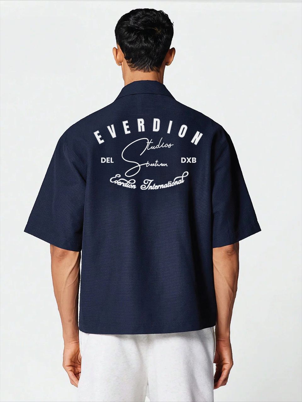 A man wearing a navy blue, oversized, half-sleeve bowling shirt with a crew neckline, featuring a small signature print on the front.