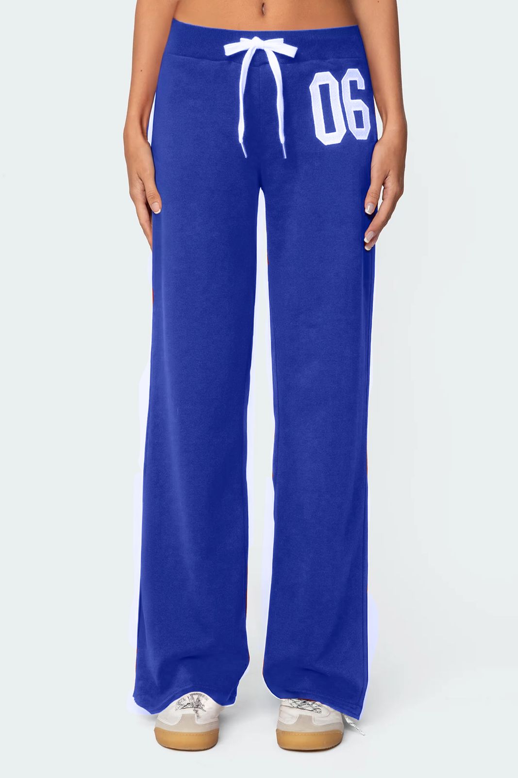 A female model wearing a white tank top and blue lowrise trackpants with the number '06' printed on the left leg.