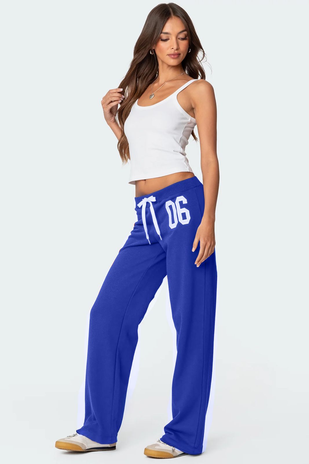 A female model wearing a white tank top and blue lowrise trackpants with the number '06' printed on the left leg.