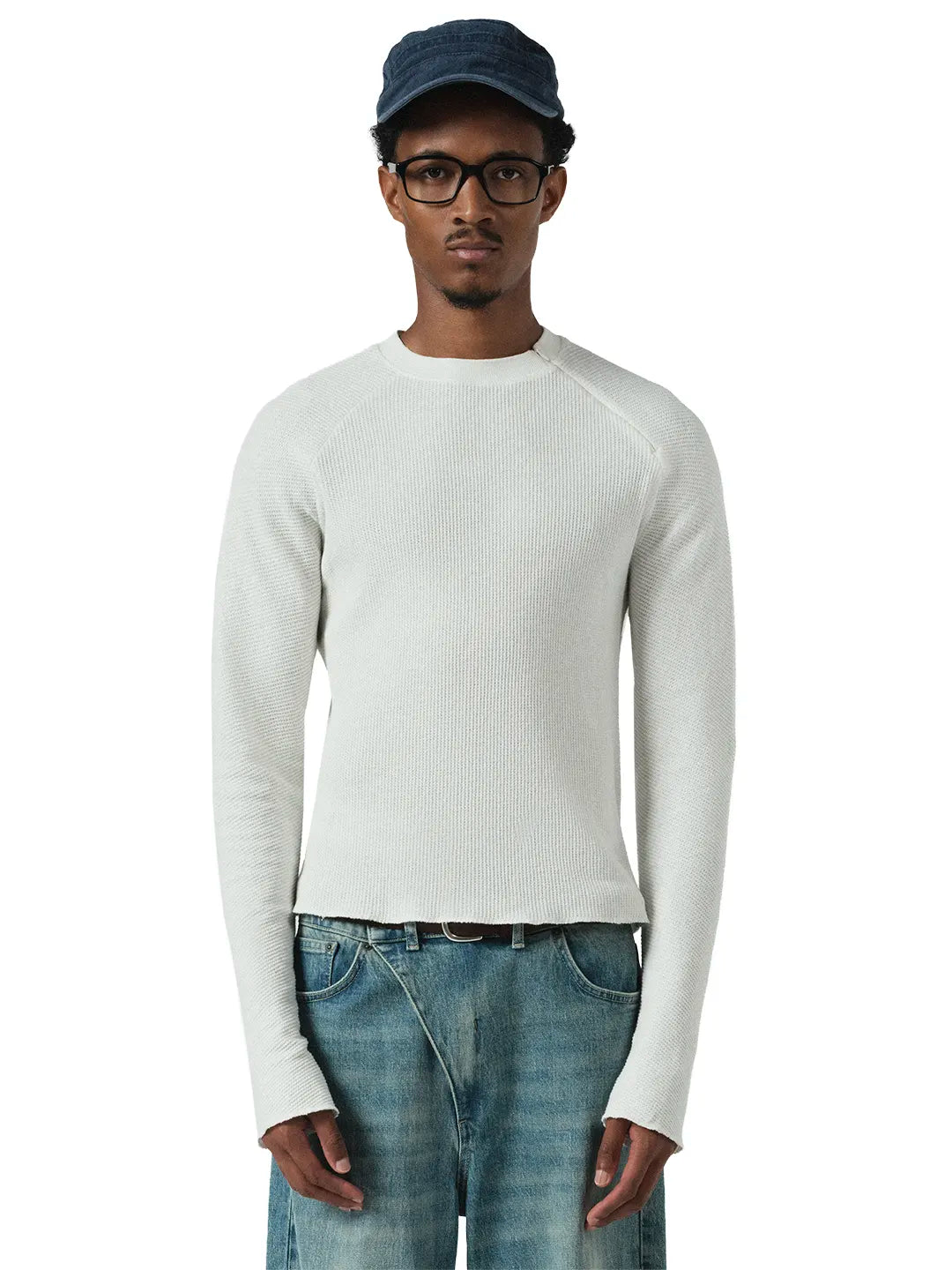 A man wearing a white long-sleeve knit sweater, blue jeans, a dark blue cap, and glasses, standing against a plain white background.