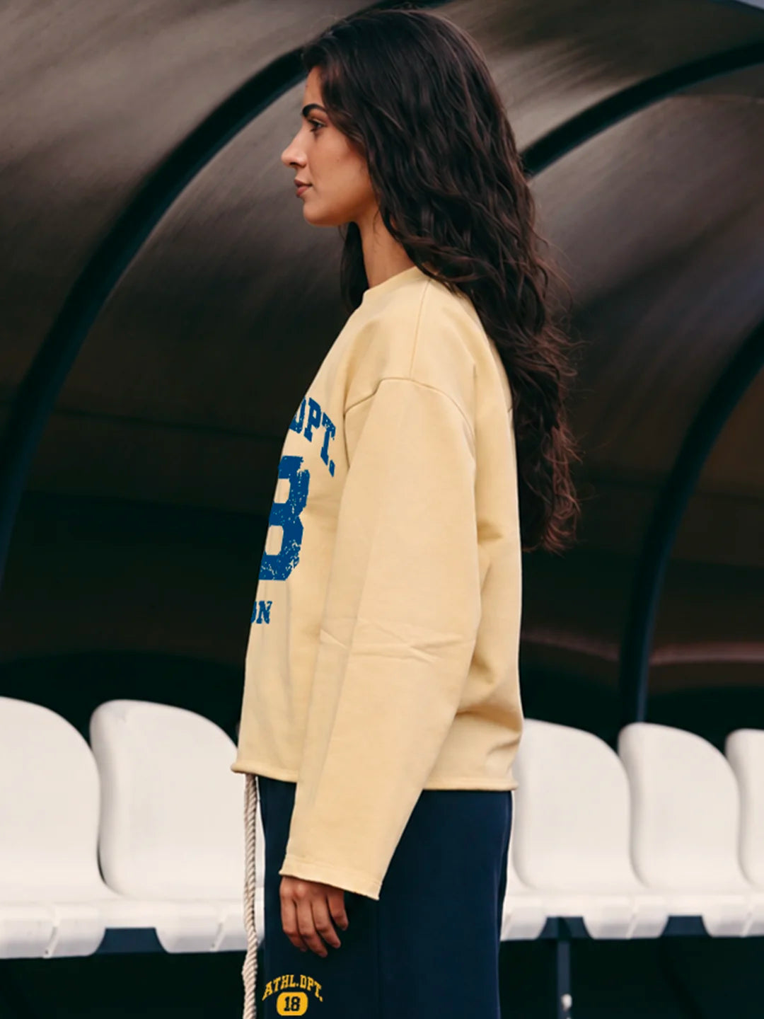 A woman wearing a cream-colored sweatshirt with blue varsity lettering and dark navy pants, standing in front of a dark arched structure with white stadium seats in the background.