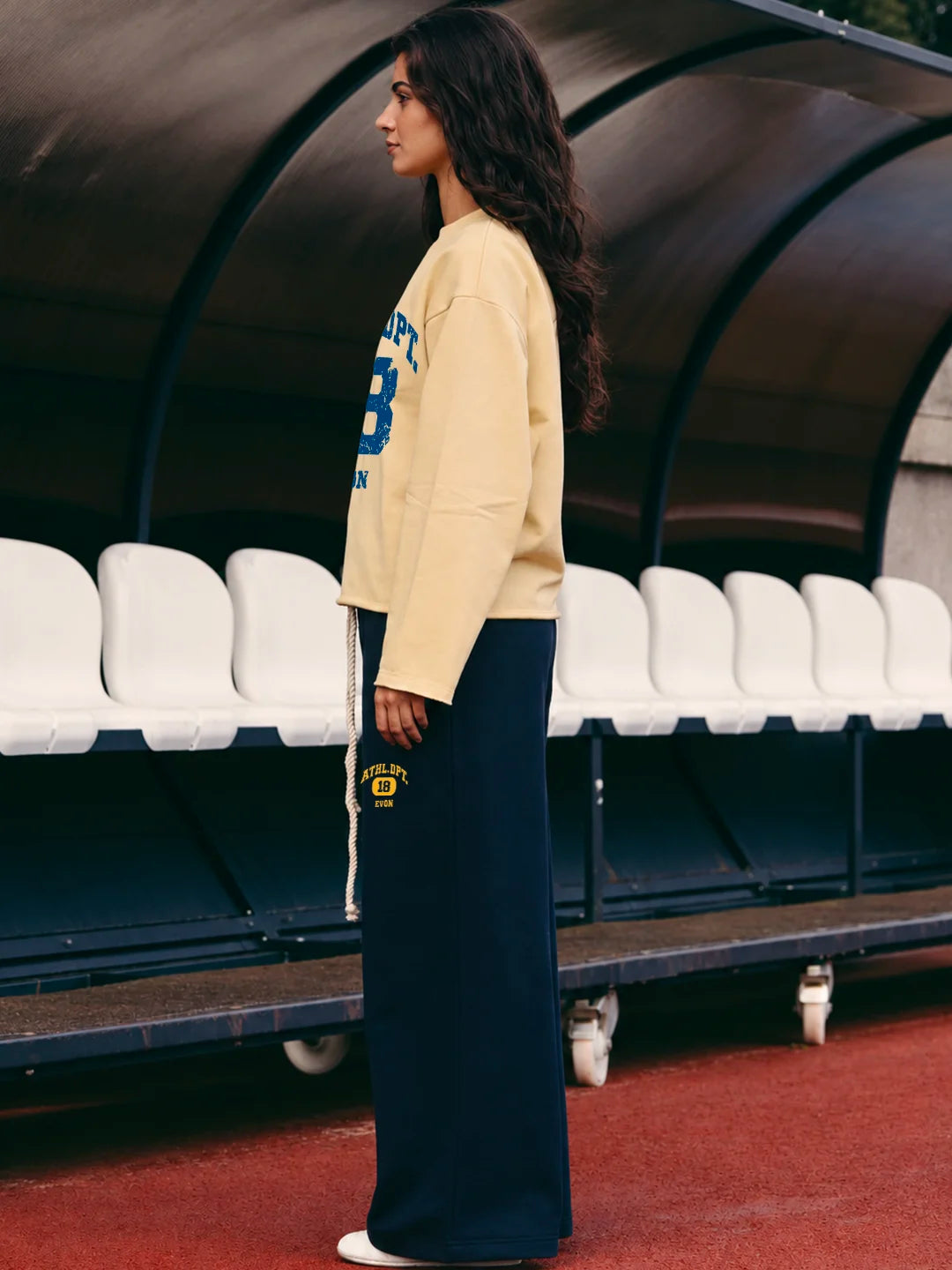 A woman wearing a cream tshirt with blue lettering and navy wide-leg sweatpants with a yellow crest patch.