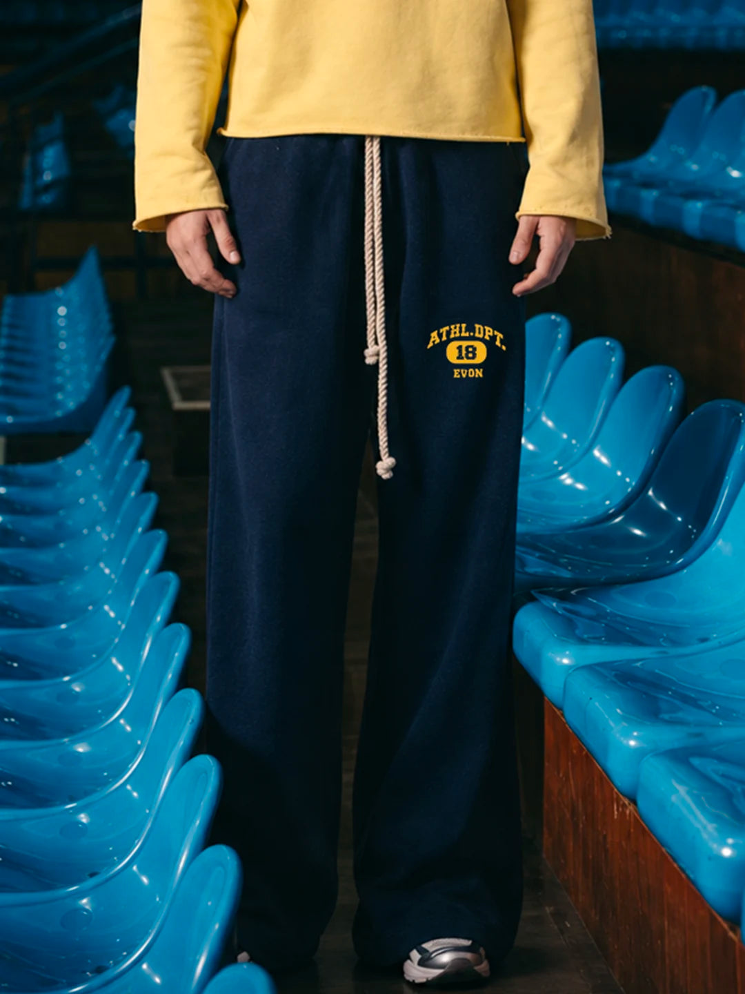 A person wearing navy wide-leg sweatpants with a yellow and white drawstring and 'AUSLIFT 18 EVON' embroidery, paired with a yellow sweatshirt, standing between rows of blue stadium seats.