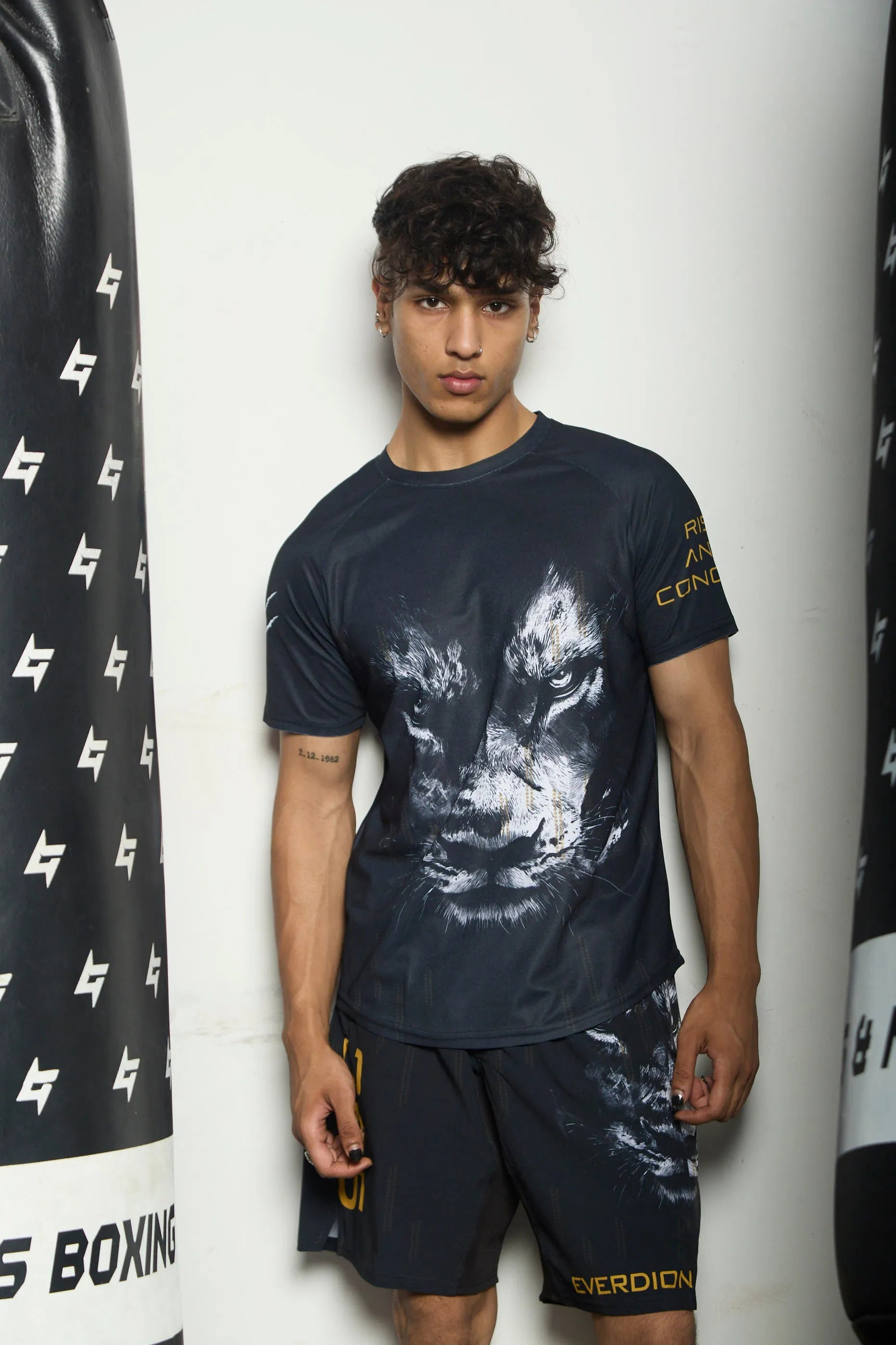 Person wearing a dark t-shirt and shorts with a brand logo, standing against a white wall.