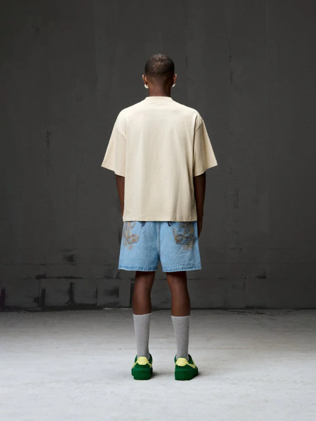 Person wearing a beige t-shirt, blue shorts, and green shoes against a grey background