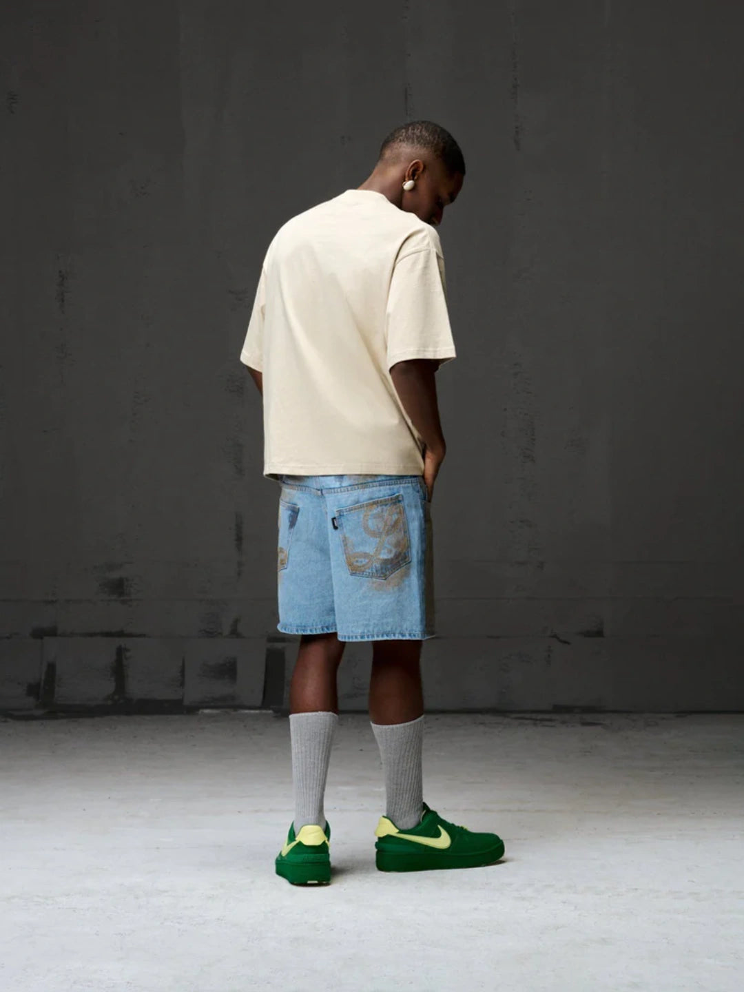 Person wearing a beige t-shirt, blue shorts, grey socks, and green sneakers in a minimalistic setting.