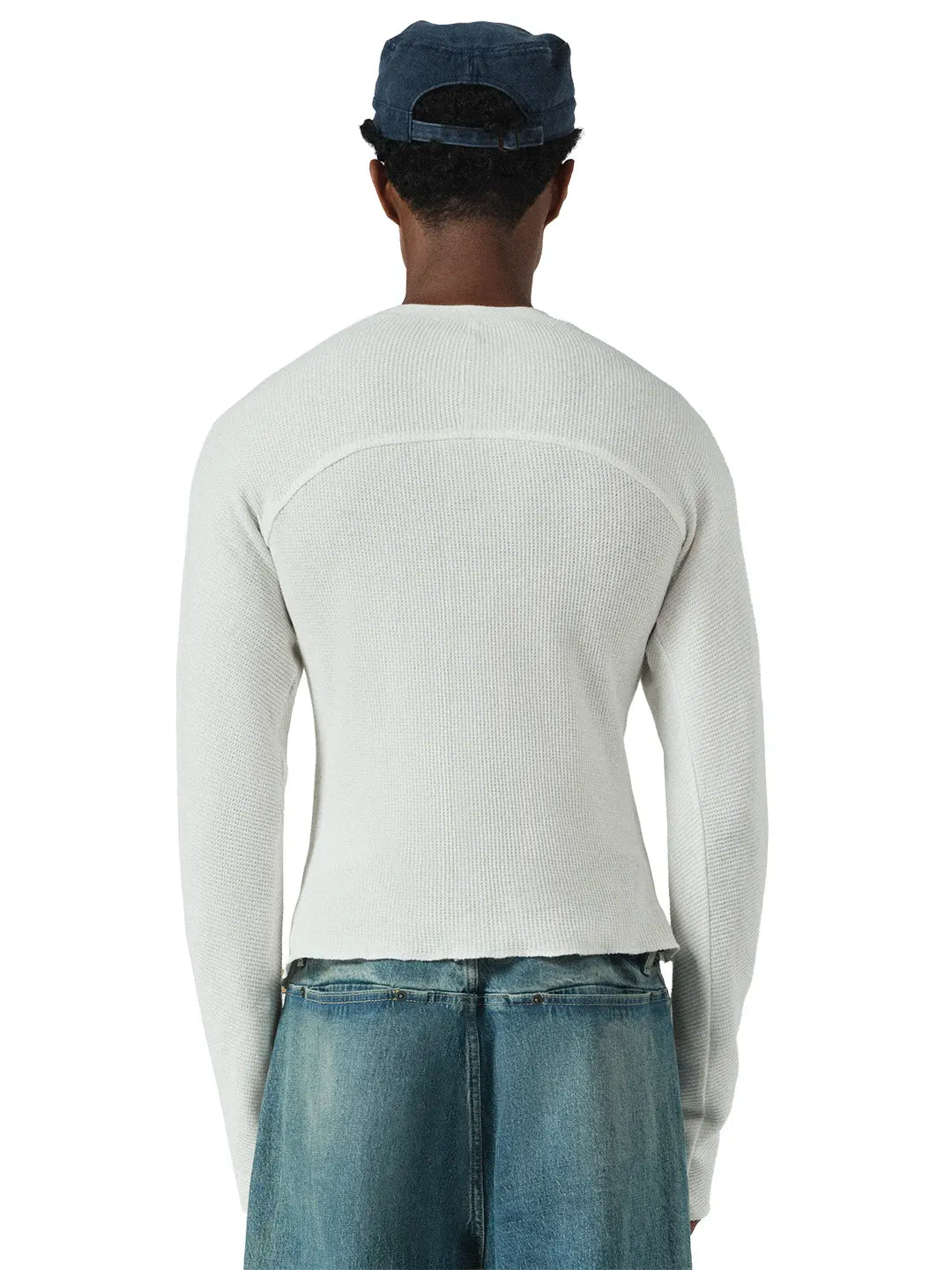 A model wearing a light grey ribbed knit sweater, blue jeans, and a dark blue baseball cap against a plain white background.