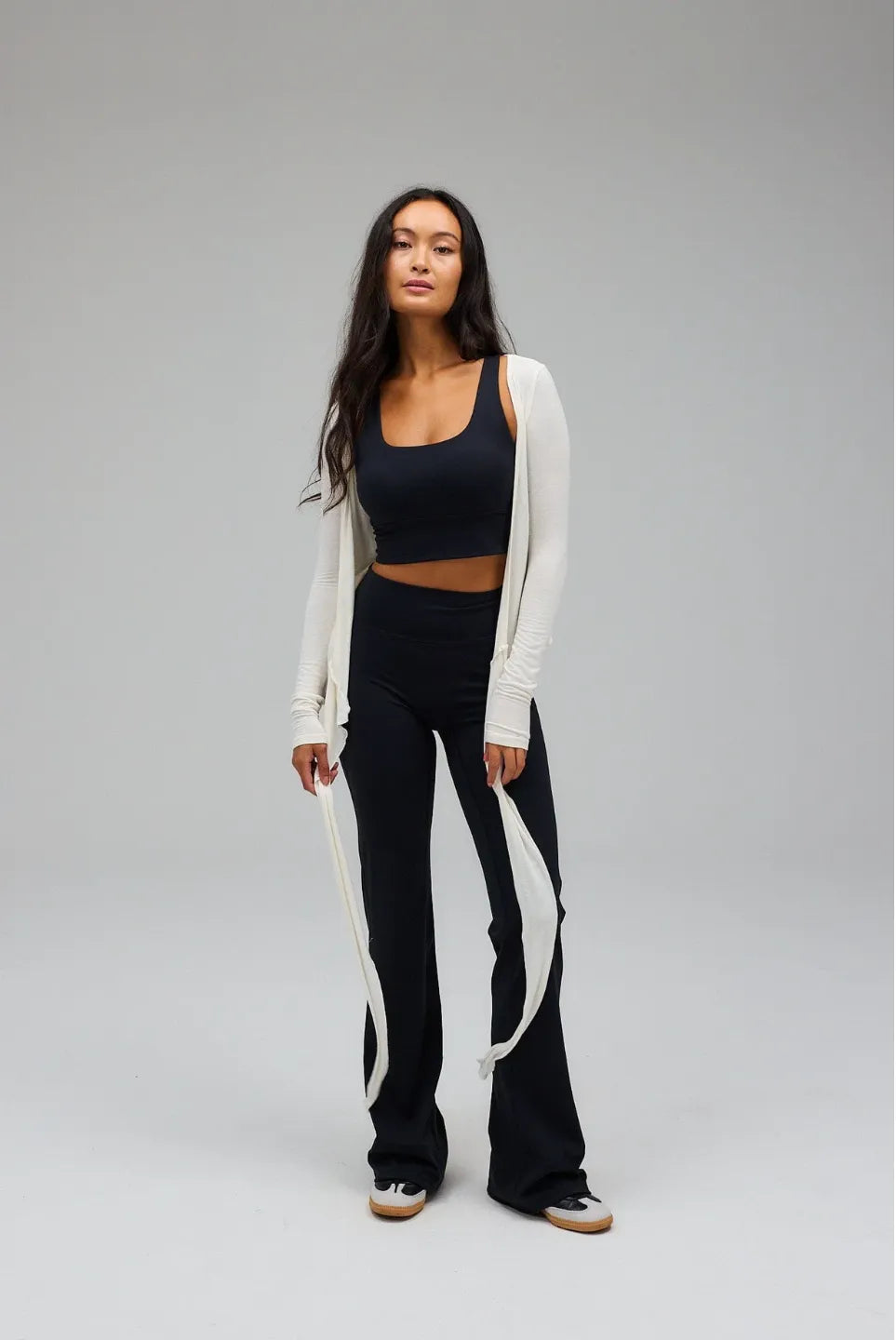 Woman wearing a black crop top, white cardigan, and black pants with white stripes on a grey background