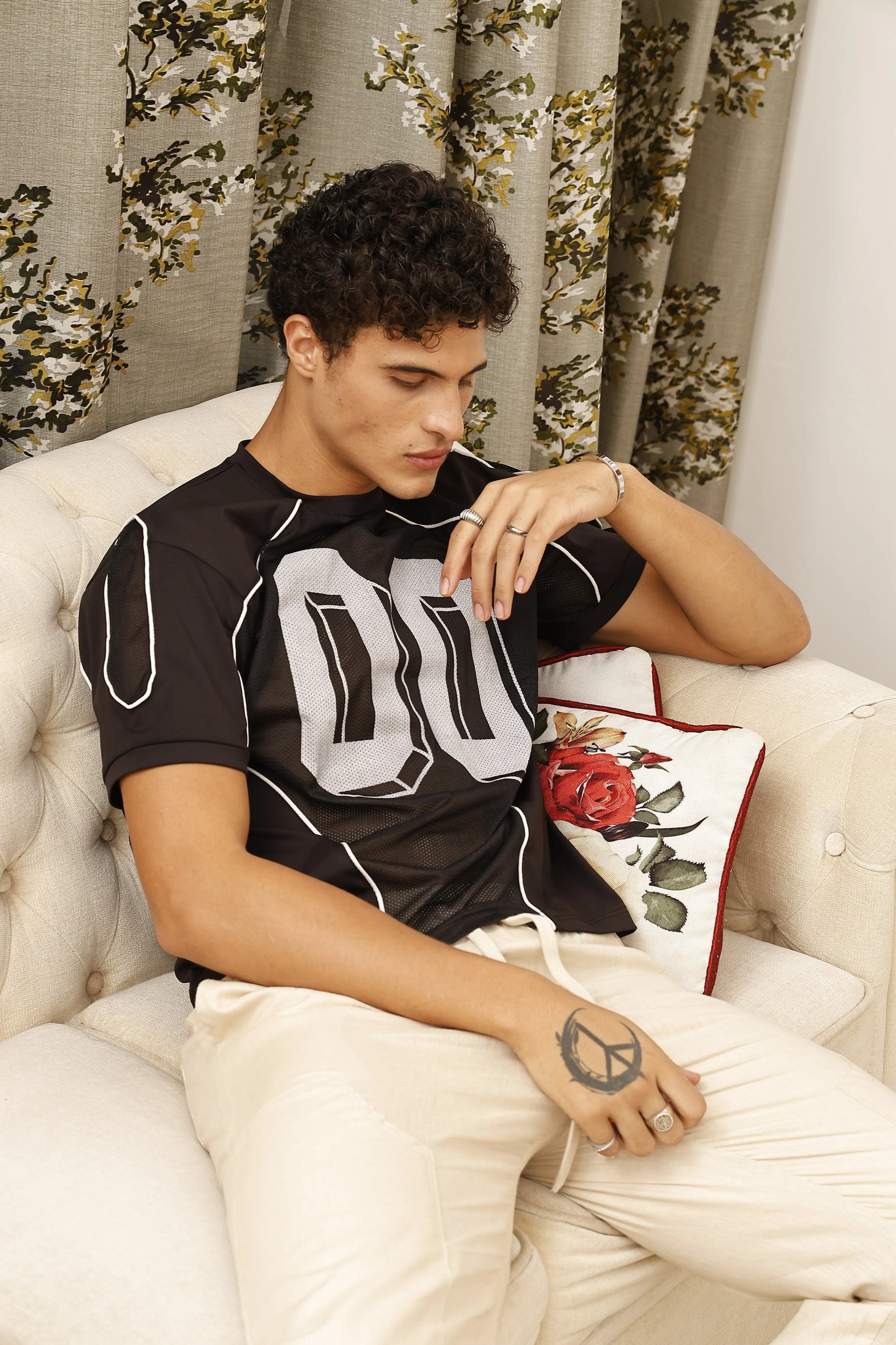 Man sitting on a white couch wearing a black shirt with a floral-patterned curtain in the background.