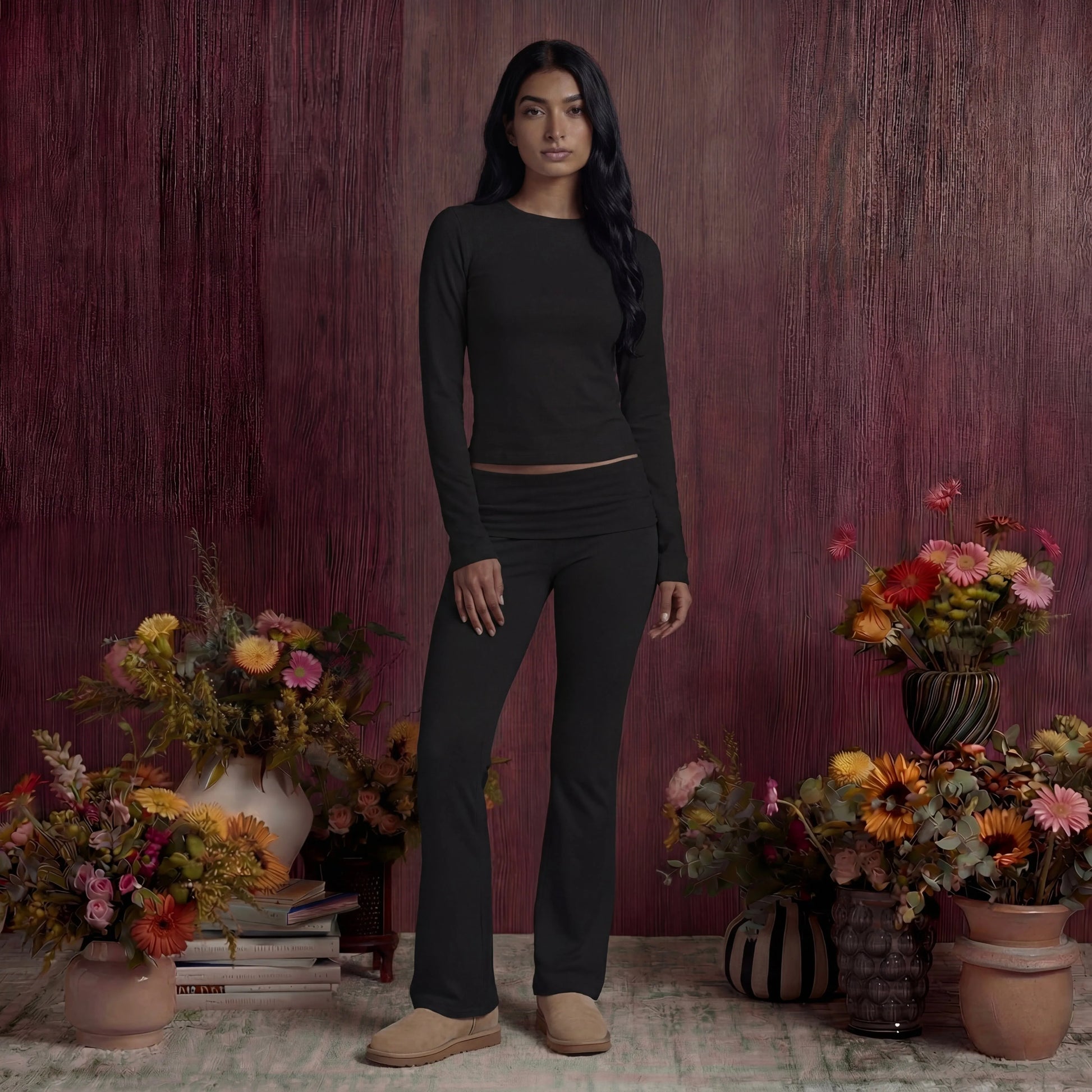 Woman in black cotton fold over pants and top standing among floral arrangements against a maroon wall.