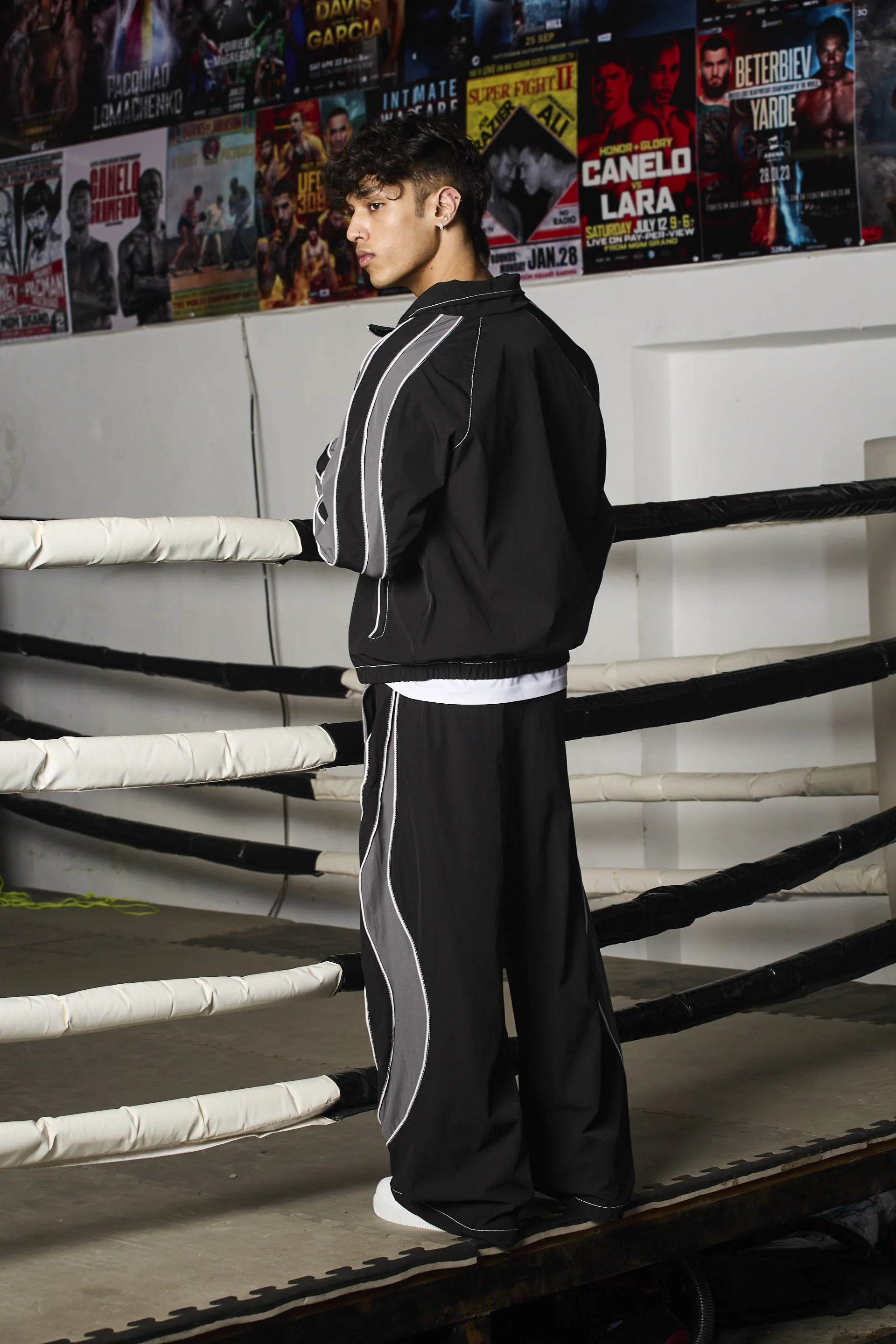 Person in a black tracksuit standing in a boxing ring with promotional posters in the background