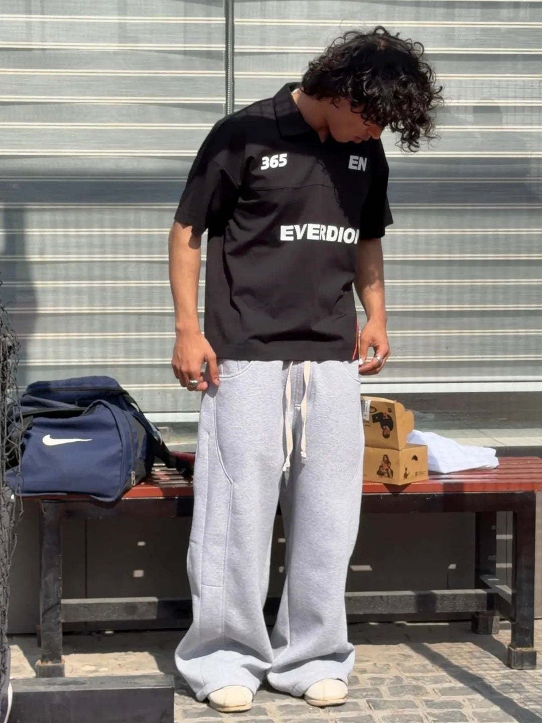 Person wearing a black 'EVERDION' shirt and grey pants standing on a bench with a striped background.