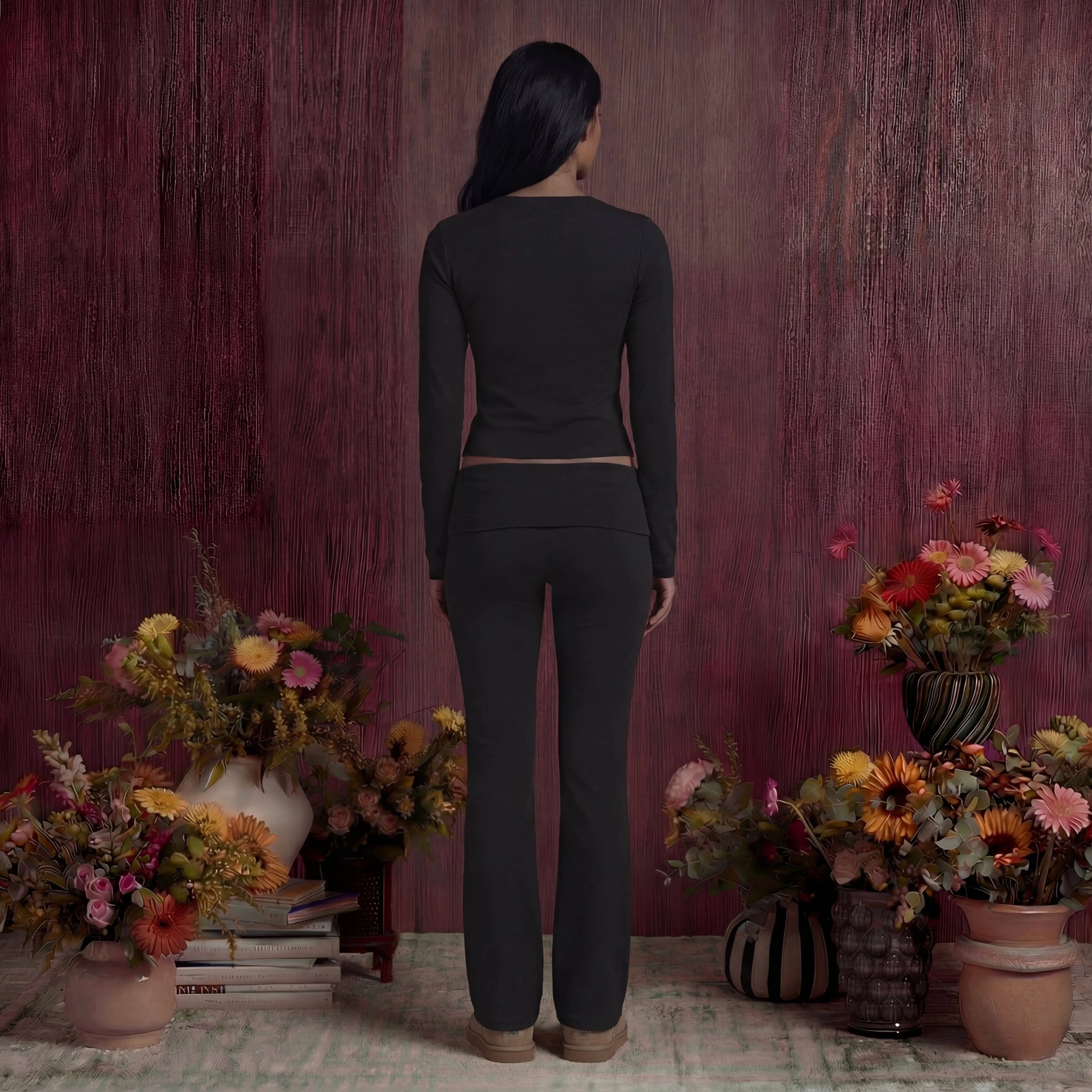 Woman in black cotton fold over pants and top standing in a room with floral arrangements and a textured wall.