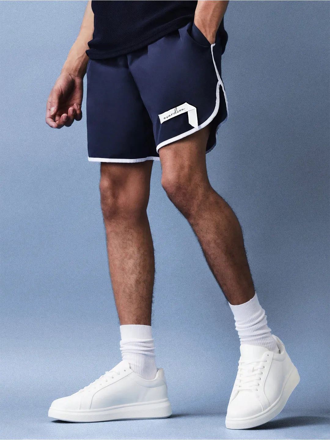Person wearing navy shorts with a white logo, white socks, and white sneakers on a light blue background