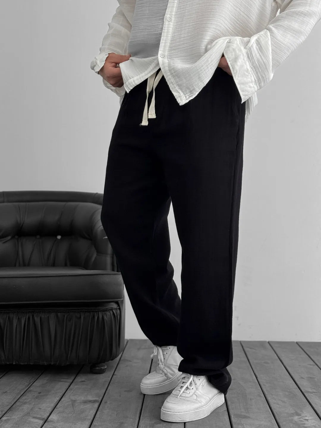 Person wearing a white oversized shirt and black pants standing in a room with a black chair.