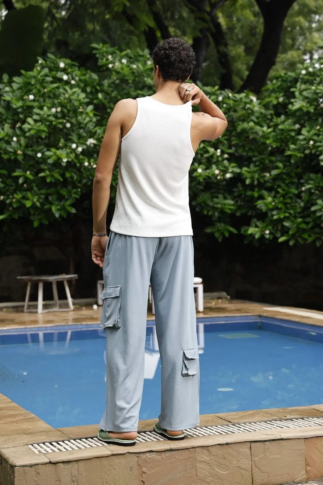 Person standing by a pool wearing a white tank top and light grey cargo pants, with greenery in the background.