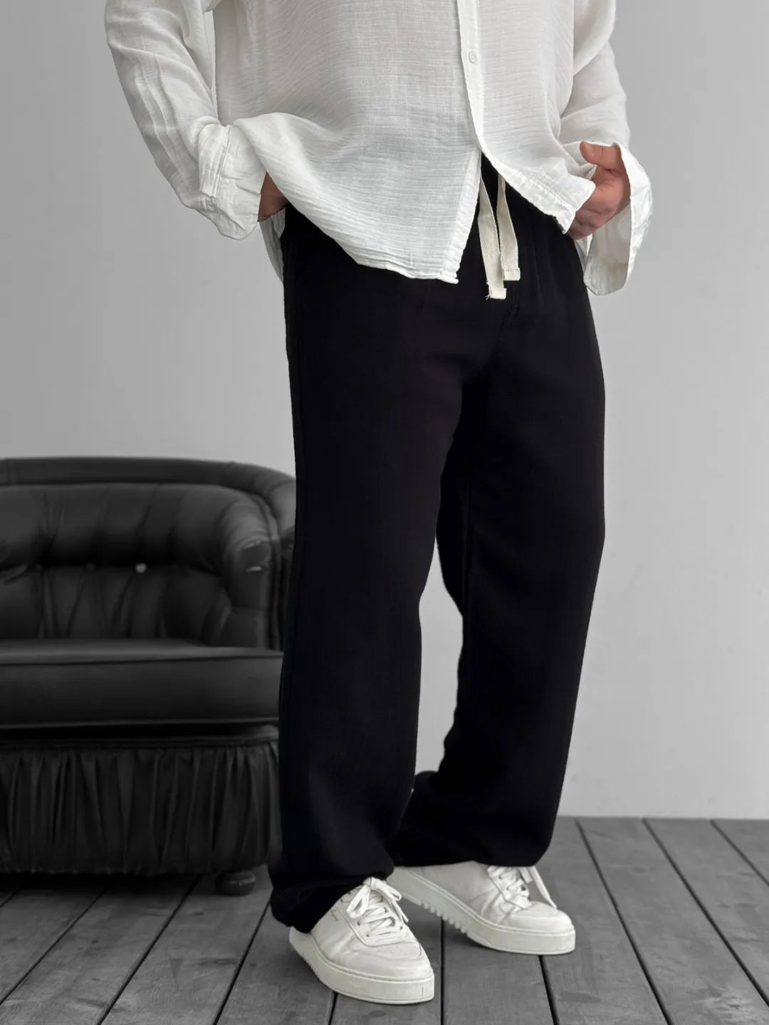 Person wearing a white shirt, black pants, and white sneakers in a minimalistic room.
