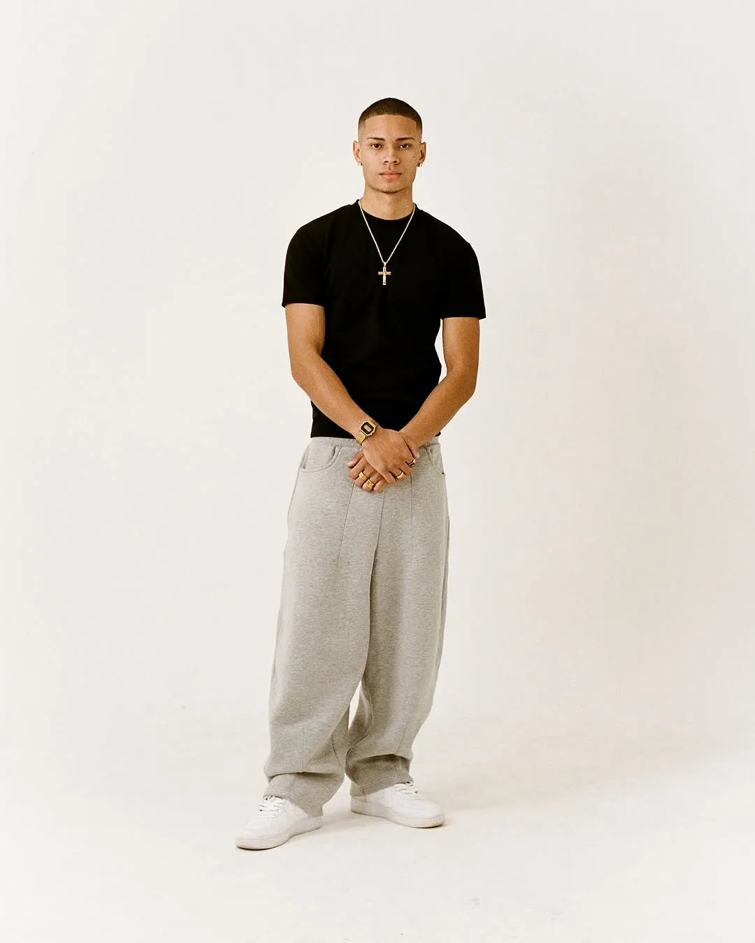 Man wearing a black fitted t-shirt, grey pants, and white shoes on a white background