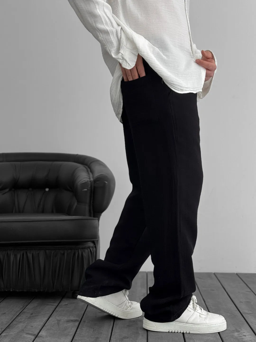 Person wearing a white shirt, black pants, and white sneakers in a minimalistic room.